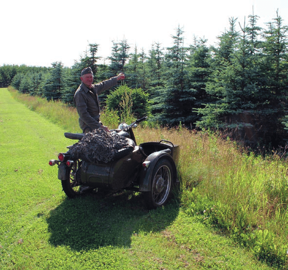 Mand står ved gammel motorcykel og fremviser området med skov og sø
